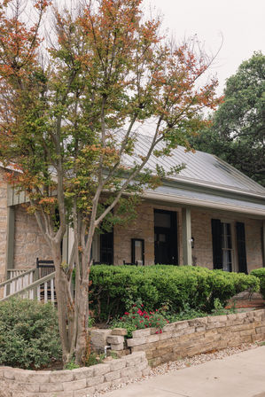 Cozy stone house with metal roof and covered front porch, autumn tree with orange-green leaves, trimmed boxwood hedge and red flowers behind a low stacked-stone retaining wall.