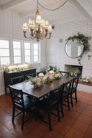 Sunlit modern farmhouse dining room with a long dark wooden table and eight black chairs, pastel floral centerpieces, ornate chandelier, white shiplap walls and round mirror above a white brick fireplace on terracotta tile floor.