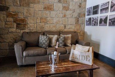 Cozy rustic bridal lounge with a brown sofa against a stone wall, patterned throw pillows, wooden coffee table holding a chilled bottle of champagne in an ice bucket and a tote bag labeled BRIDE.