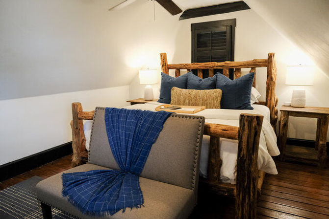 Cozy attic bedroom with rustic log bed, navy pillows and a woven lumbar pillow, bedside lamps, dark wooden floors and a gray bench draped with a blue throw.