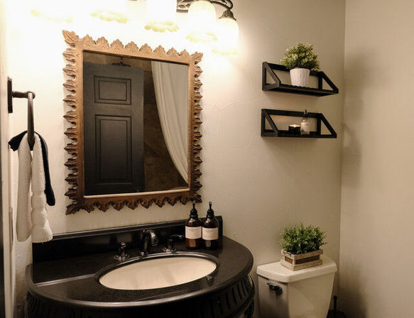 Cozy chic bathroom with ornate wood-framed mirror over a black oval vanity and sink, black fixtures, amber soap dispensers, floating black shelves with potted plants and a candle, towel on rail, and toilet topped with a small planter.