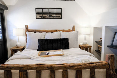 Cozy rustic bedroom with reclaimed log bed frame, white linens and striped pillows, black lumbar cushion, bedside lamps, wooden nightstands and a tray with remote on the bed.
