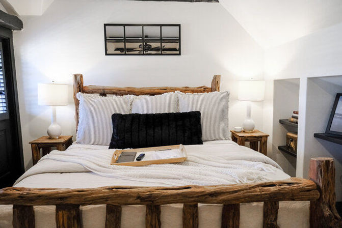 Cozy rustic bedroom with reclaimed log bed frame, white linens and striped pillows, black lumbar cushion, bedside lamps, wooden nightstands and a tray with remote on the bed.