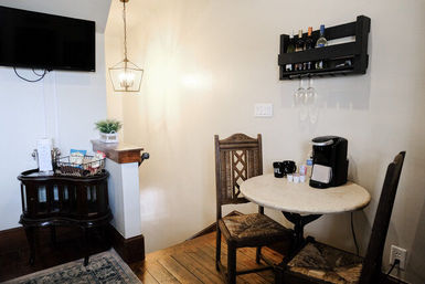 Cozy breakfast nook in a bright room with a round bistro table, two carved wooden chairs, single-serve coffee maker and mugs, wall-mounted wine rack with hanging glasses, geometric pendant light, small TV and snack basket on a vintage side table over hardwood floors.