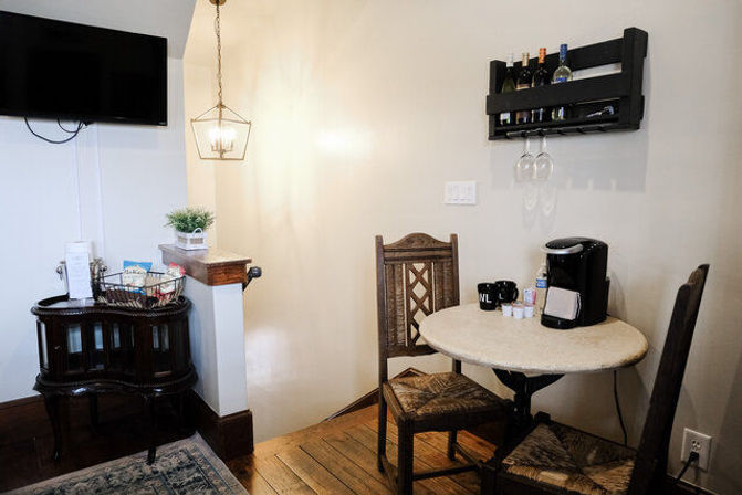Cozy breakfast nook in a bright room with a round bistro table, two carved wooden chairs, single-serve coffee maker and mugs, wall-mounted wine rack with hanging glasses, geometric pendant light, small TV and snack basket on a vintage side table over hardwood floors.