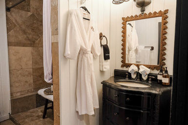 Cozy spa-style bathroom interior with tiled walk-in shower, white waffle robe hanging on shiplap wall, dark curved vanity with marble sink and ornate gold mirror