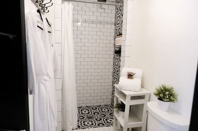 Spa-like modern white bathroom with subway-tile shower, black-and-white patterned floor tiles, white shower curtain and hanging robes, stacked towels on a small wooden shelf, and a potted plant on the toilet tank.