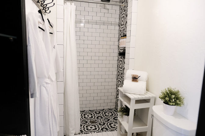 Spa-like modern white bathroom with subway-tile shower, black-and-white patterned floor tiles, white shower curtain and hanging robes, stacked towels on a small wooden shelf, and a potted plant on the toilet tank.