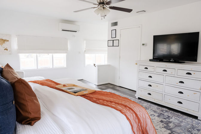 Sunny modern master bedroom with white walls and windows, king bed dressed in white linens with rust-orange throw and pillows, wooden breakfast tray, distressed white dresser with flat-screen TV, ceiling fan and wall-mounted AC.