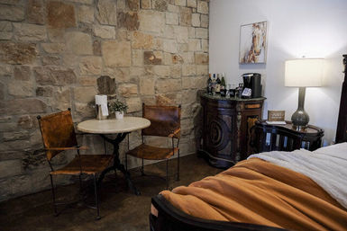Cozy rustic bedroom corner with exposed stone wall, small round bistro table and two leather chairs, antique cabinet with coffee setup, bedside lamp and bed with a warm orange throw.