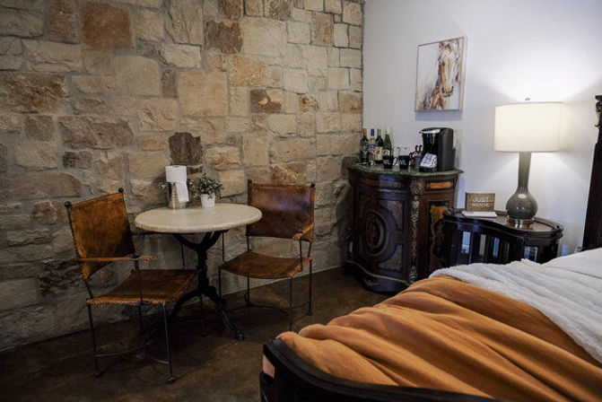 Cozy rustic bedroom corner with exposed stone wall, small round bistro table and two leather chairs, antique cabinet with coffee setup, bedside lamp and bed with a warm orange throw.