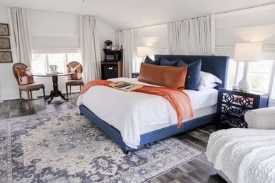 Bright modern farmhouse bedroom interior with a blue upholstered bed, white linens and rust-orange throw, patterned area rug, round bistro table by windows, bedside lamps and hardwood floors.