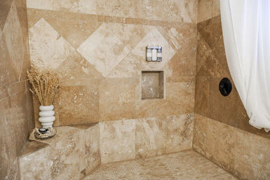 Beige travertine tiled walk-in shower with diamond-pattern accent tiles, built-in corner bench holding a white vase of dried grasses and soap dish, recessed niche with bottles, black shower control and a white shower curtain.