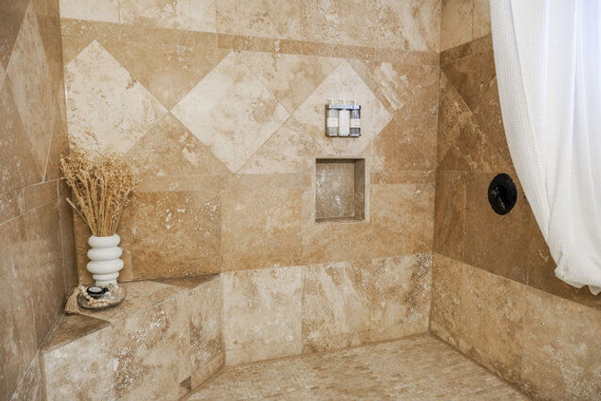 Beige travertine tiled walk-in shower with diamond-pattern accent tiles, built-in corner bench holding a white vase of dried grasses and soap dish, recessed niche with bottles, black shower control and a white shower curtain.