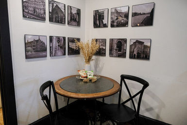 Cozy cafe corner with a small round wooden table and two black cross-back chairs, dried wheat centerpiece in a vase and snack packets on a plate, framed black-and-white photos on a white gallery wall.