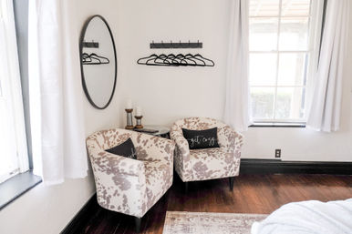Sunlit bedroom corner with two cowhide-pattern accent chairs and 'get cozy' pillows, round wall mirror, black wall-mounted coat rack with hangers, white curtains and dark hardwood floor.