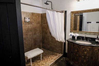 Rustic bathroom featuring a tiled walk-in shower with white curtain and small bench, wall-mounted rain showerhead, and an ornate carved wooden vanity with black countertop and gilded mirror.