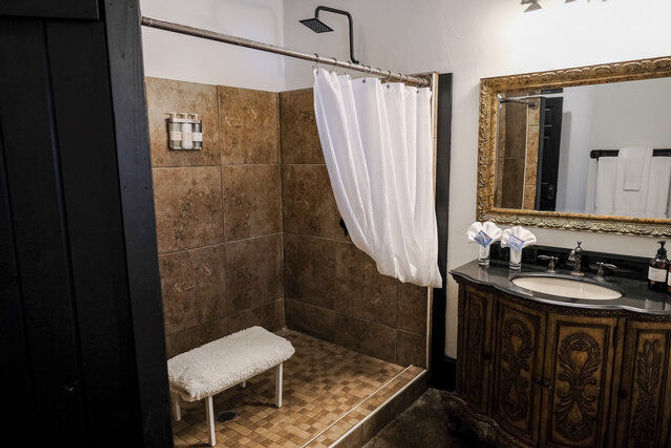 Rustic bathroom featuring a tiled walk-in shower with white curtain and small bench, wall-mounted rain showerhead, and an ornate carved wooden vanity with black countertop and gilded mirror.