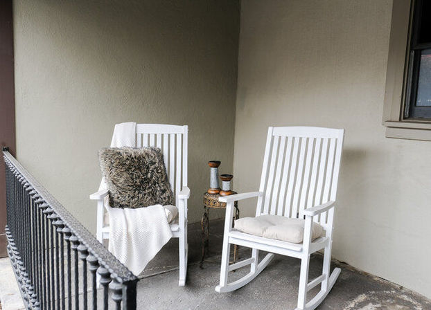 Cozy front porch scene with two white wooden rocking chairs, one draped with a white throw and faux fur pillow, small round side table with candle holders, neutral stucco wall and iron railing.