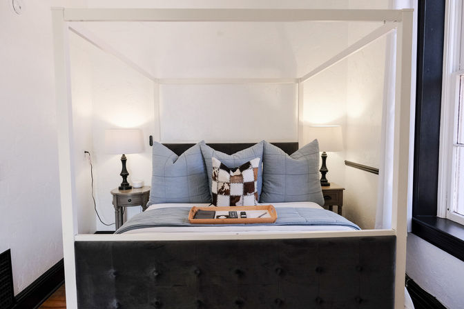 Sunlit cozy bedroom with a white four-post canopy bed, gray tufted headboard, blue-gray pillows and quilt, patterned accent pillow, wooden nightstands and lamps by a window.