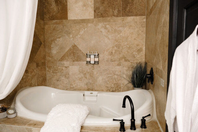 Spa-like bathroom with white soaking tub set into beige travertine tile surround, black matte faucet, wall-mounted soap dispensers, white towel and shower curtain.