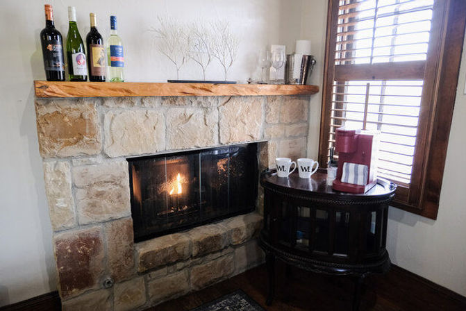 Cozy corner with lit stone fireplace and wooden mantel, wine bottles and twig decor above, round dark-wood side table with red single-serve coffee maker and two mugs by a shuttered window.