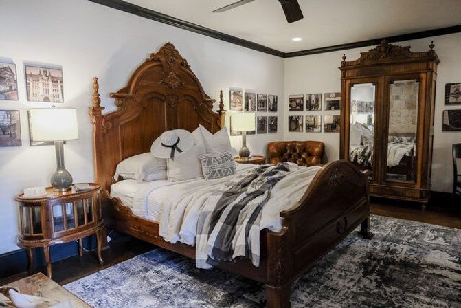Cozy vintage bedroom interior with ornate carved wooden bed, striped throw and pillows, mirrored antique armoire, tufted leather chair, bedside lamps, framed wall photos and textured area rug.