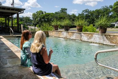 Two women relaxing at the edge of a sunlit outdoor stone pool, feet in clear turquoise water, one holding a drink, with a low stone waterfall, large potted plants on the pool ledge and trees beyond.