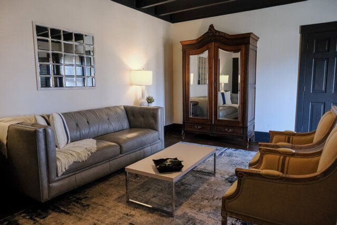 Cozy living room interior with gray tufted leather sofa, two beige upholstered armchairs, low white coffee table on a patterned rug, antique wooden mirrored armoire, wall mirror and table lamp — stylish modern-meets-vintage decor.