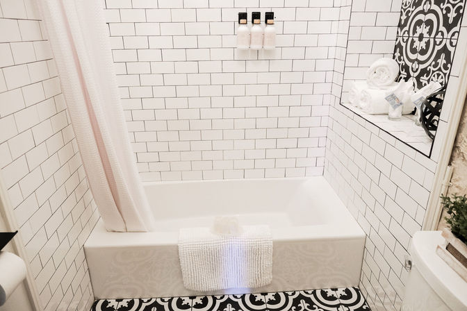 Bright bathroom with white subway-tiled bathtub, black-and-white patterned floor tile, shower curtain, wall-mounted soap dispensers and a recessed shelf holding rolled towels and toiletries.