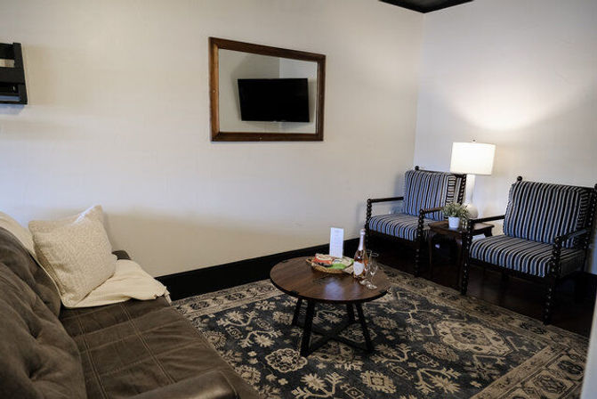 Cozy lounge interior with brown leather sofa, two striped armchairs, wooden coffee table with wine and glasses, patterned area rug and mirror reflecting a wall-mounted TV