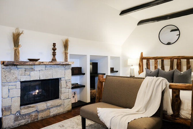 Cozy rustic mountain cabin bedroom interior with stone fireplace, reclaimed log bed, tufted bench draped with a white throw, built-in shelves, round wall mirror and warm bedside lamp.