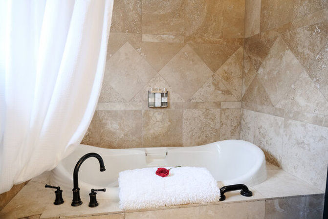 Spa-like bathroom with a white alcove bathtub set in beige marble tile, black faucet and handles, white shower curtain, and a folded fluffy white towel topped with a red rose.
