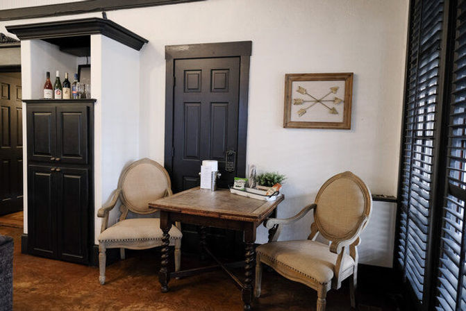 Cozy interior seating nook with a vintage wooden table flanked by two beige upholstered round-back chairs, black paneled door and shutters, cabinet with wine bottles, framed arrow art and a small plant.