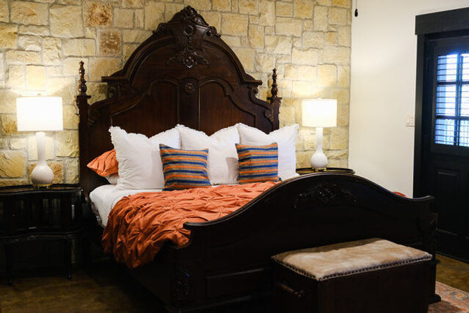 Cozy rustic bedroom with ornate carved dark-wood bed against a stone accent wall, orange duvet, striped accent pillows, white shams and matching bedside lamps.