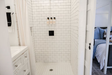 Bright white subway-tile shower with hexagon floor tiles, black square valve and three wall-mounted dispensers, white vanity and curtain, view into a cozy bedroom with blue bedding.