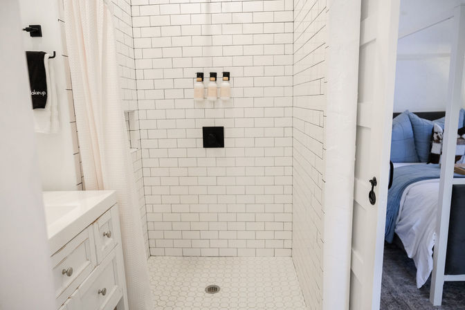 Bright white subway-tile shower with hexagon floor tiles, black square valve and three wall-mounted dispensers, white vanity and curtain, view into a cozy bedroom with blue bedding.