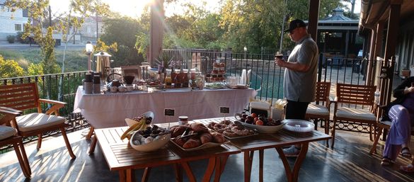 Sunlit outdoor patio breakfast buffet on a wooden deck overlooking water and trees, with a table of coffee urns, juices, stacked pastries, croissants and fruit bowls, and a man in a cap checking his phone near poolside seating.