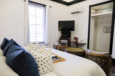 Cozy bright bedroom interior with large window, plush navy and gold-dot pillows on a white bed, wall-mounted TV, mini-fridge and coffee station, round wicker table and two cushioned chairs.