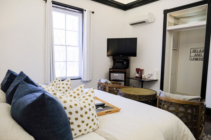 Cozy bright bedroom interior with large window, plush navy and gold-dot pillows on a white bed, wall-mounted TV, mini-fridge and coffee station, round wicker table and two cushioned chairs.