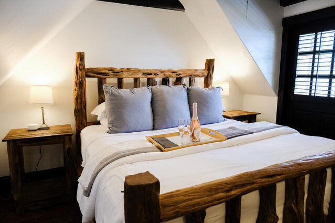 Cozy rustic attic bedroom with a reclaimed wood bed, white linens and gray pillows, bedside lamps and a tray holding a bottle of rosé and two glasses by a shuttered window.