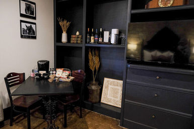 Cozy dining nook with a black table and two wooden chairs, a coffee maker and snacks on the table, dark built-in shelving holding wine bottles and decorative wheat, framed horse photos on the wall, and a flat-screen TV on a nearby dresser.