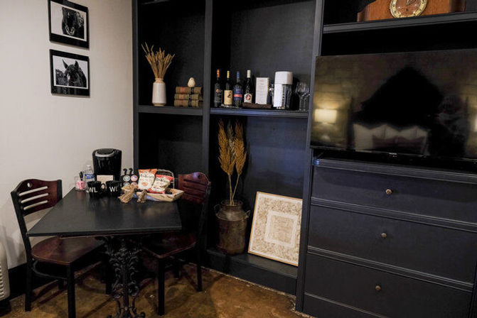 Cozy dining nook with a black table and two wooden chairs, a coffee maker and snacks on the table, dark built-in shelving holding wine bottles and decorative wheat, framed horse photos on the wall, and a flat-screen TV on a nearby dresser.