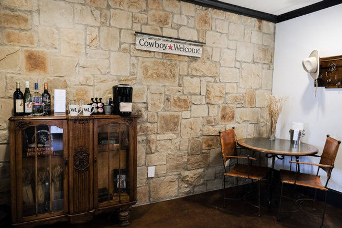 Rustic western cafe corner with stone wall and 'Cowboy Welcome' sign, vintage wooden cabinet with wine bottles and coffee station, round metal table and leather chairs.