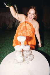 Person in a bright orange dress pouring bubbly into a stacked coupe glass tower outdoors at night, foam overflowing onto a white tablecloth, smiling energetically.