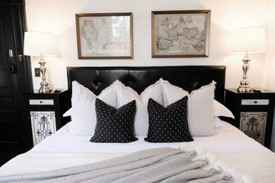 Cozy stylish bedroom with black tufted headboard, striped and cross-pattern pillows, silver bedside lamps, framed vintage maps, and a soft gray throw.