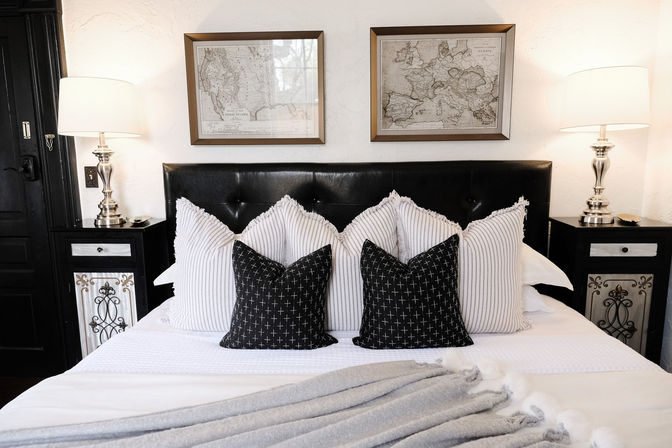 Cozy stylish bedroom with black tufted headboard, striped and cross-pattern pillows, silver bedside lamps, framed vintage maps, and a soft gray throw.