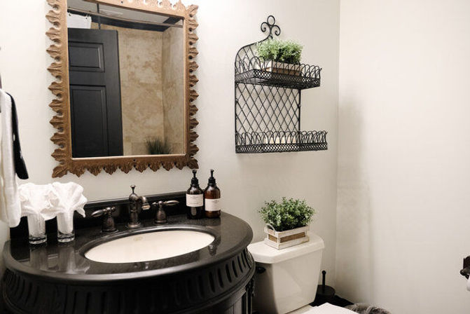 Cozy farmhouse-style powder room with ornate wood-framed mirror, black stone vanity and oval sink, bronze faucet, wire wall shelves and potted greenery above the toilet