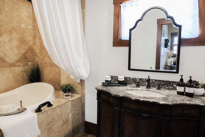 Spa-like neutral bathroom with marble-top vanity and dark wood cabinet under an arched mirror, beige-tiled soaking tub with white curtain and small potted greenery.