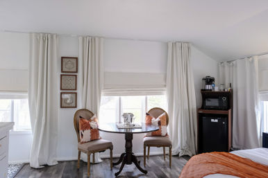Bright, cozy guest room with a round pedestal table and two vintage upholstered chairs with rust-pattern pillows, floor-to-ceiling white curtains, wood-look flooring, mini fridge and microwave coffee station, and an orange throw on the bed.